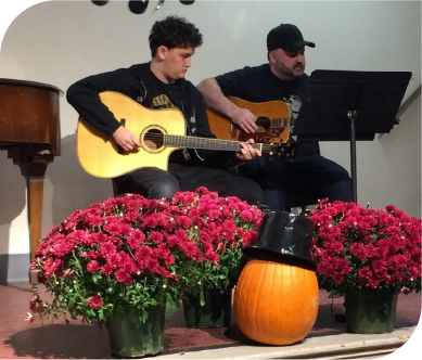 Student and instructor performing an acoustic guitar duet during a fall concert with seasonal decorations.