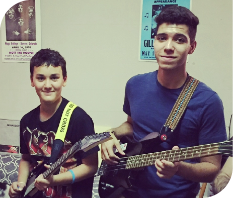 Two students smiling while practicing guitar and bass together during a music lesson.