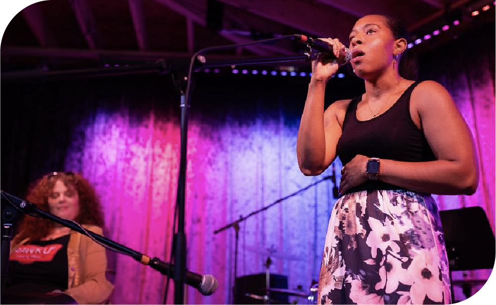 Female vocalist performing on stage with microphone under purple lighting during Rawk U School of Music concert in Philadelphia