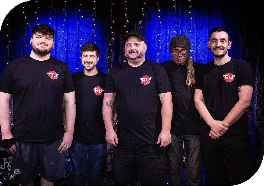 Five professional music instructors from Rawk U School of Music standing together wearing matching black shirts with RU logo against blue stage curtain backdrop in Philadelphia