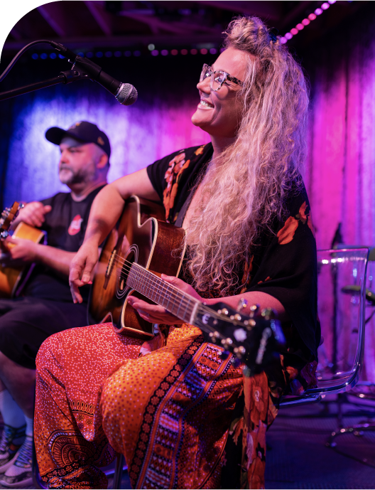 Female guitarist smiling and singing on stage during a live music performance alongside her instructor.