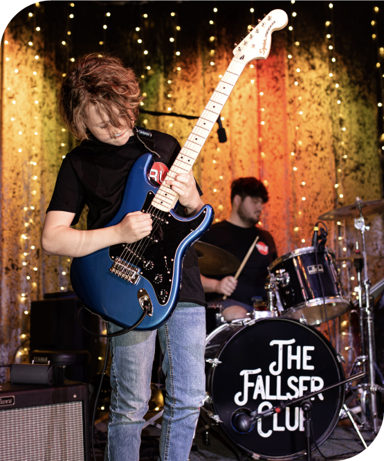 Young guitarist playing electric guitar on stage with drummer in background at a student rock band show.