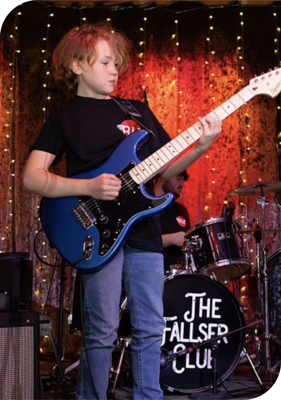 Young guitar student performing on stage with blue electric guitar in front of drum kit at Rawk U School of Music concert showcase